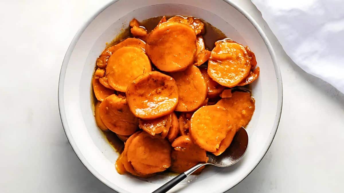 A bowl of glazed sweet potatoes, sliced and coated in a syrupy sauce. A spoon rests in the bowl, and the dish is served on a white surface with a white napkin nearby.