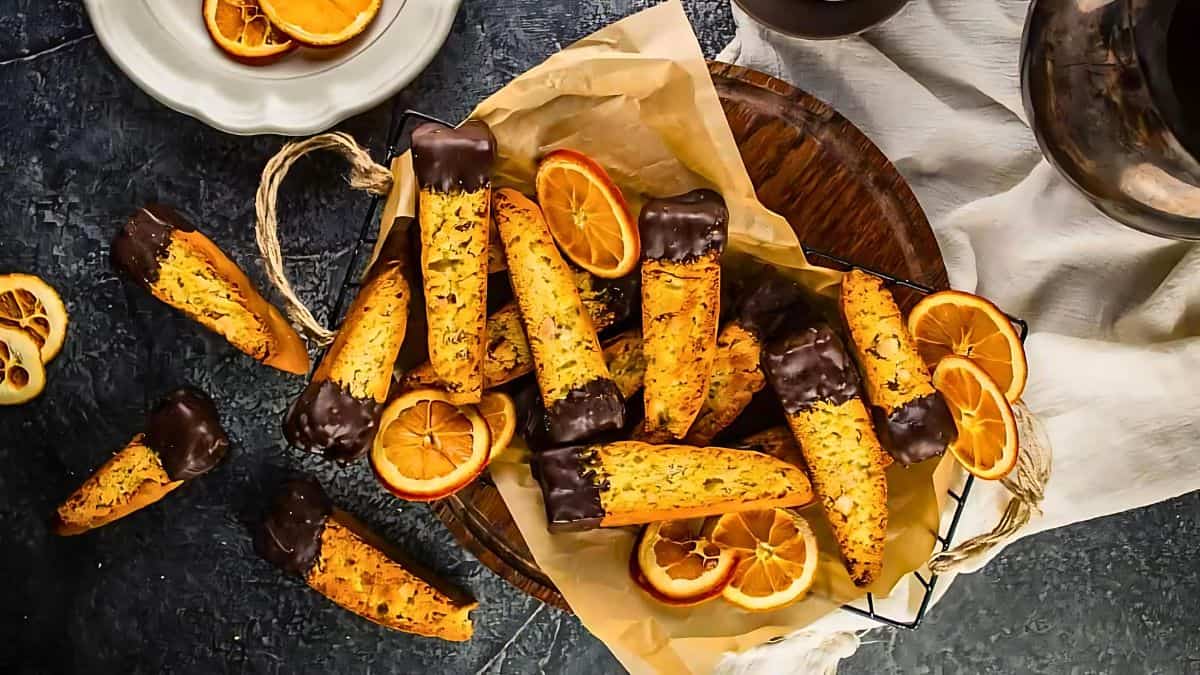 A basket filled with chocolate-dipped biscotti arranged on parchment paper, surrounded by sliced dried oranges. A plate with more dried oranges is nearby on a dark surface.