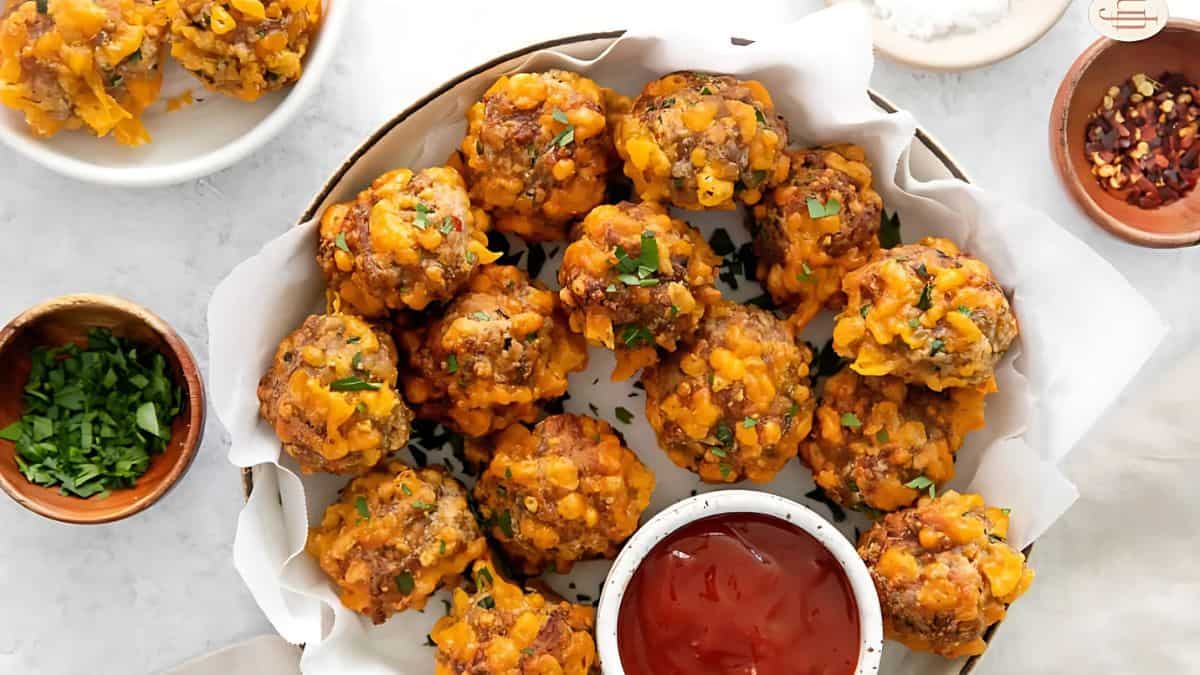 A plate of homemade sausage balls with melted cheese, garnished with chopped herbs. The plate is lined with parchment paper and accompanied by a bowl of red dipping sauce. Small bowls of herbs and chili flakes are beside the plate.