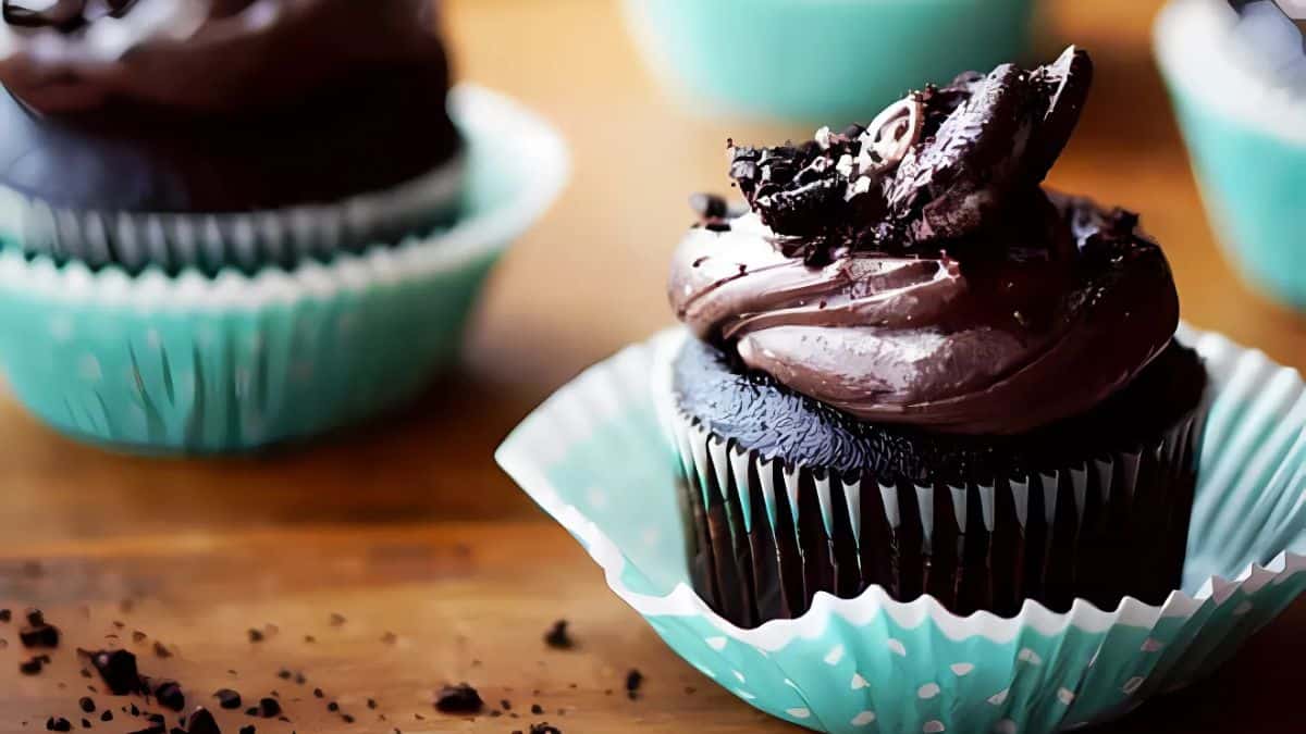 A chocolate cupcake with rich dark frosting and crumbled chocolate pieces on top, placed in a light blue cupcake liner. It's set on a wooden surface alongside other similar cupcakes in matching liners.