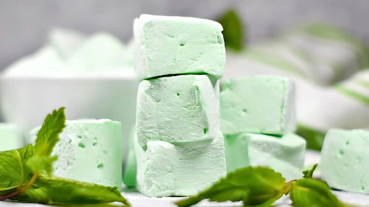A stack of four light green mint-flavored marshmallows on a white surface, surrounded by fresh mint leaves. A blurred background features a bowl of marshmallows and additional mint leaves.