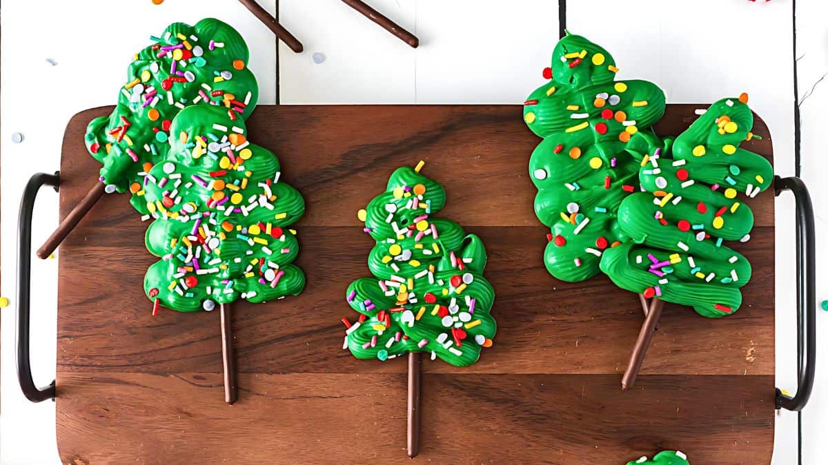 Three tree-shaped treats covered in green icing and multicolored sprinkles are placed on a wooden serving board. Each treat is on a stick, resembling a lollipop.