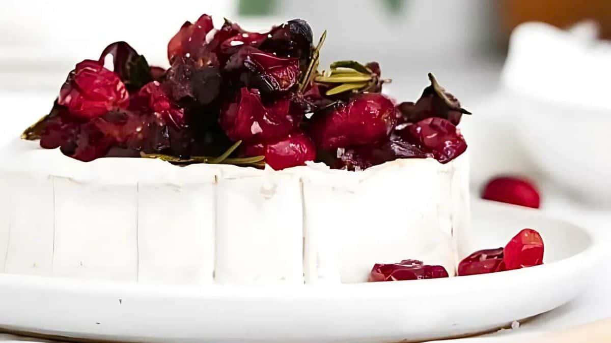 A creamy white cheese topped with a mixture of glazed cranberries and herbs sits on a white plate. The vibrant red berries contrast with the cheese, creating an appealing and festive presentation.