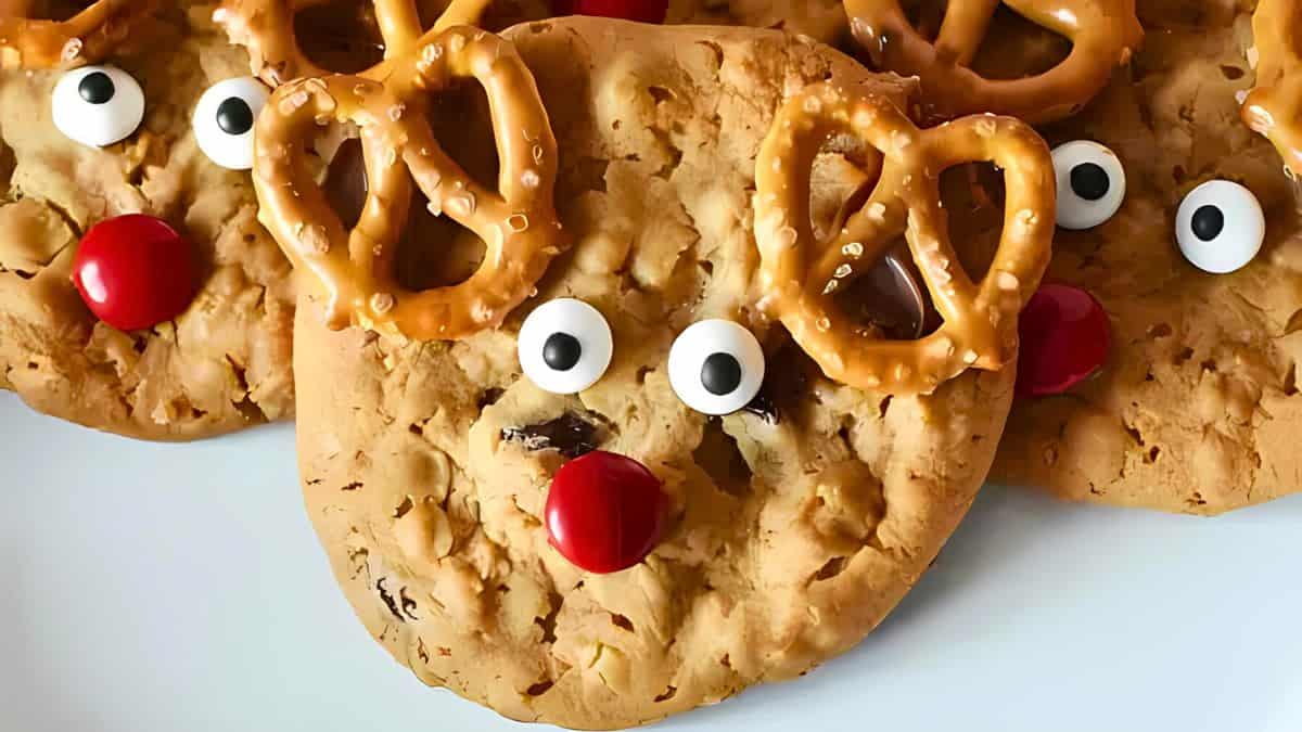 Cookies decorated to look like reindeer with pretzel antlers, candy eyes, and red noses are arranged on a plate.