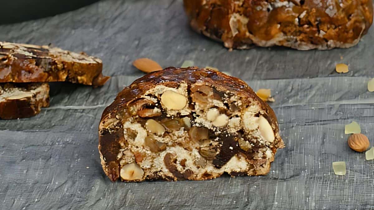 A sliced loaf of fruit and nut bread with visible almonds and other nuts on a gray surface. Another loaf and scattered almonds are in the background.