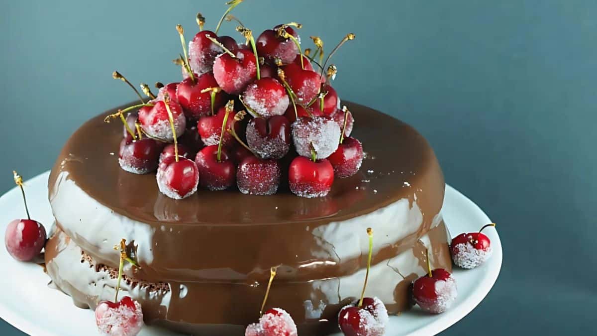 A chocolate cake topped with a mound of fresh red cherries, some of which are frosted. The cake sits on a white plate against a gray background, with a few cherries scattered around the base.