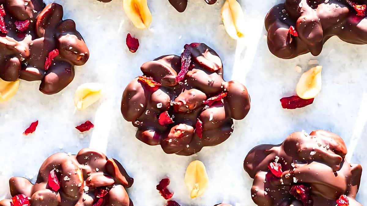 Clusters of chocolate-covered peanuts topped with sea salt and dried cranberry pieces are arranged on a parchment paper-lined surface.