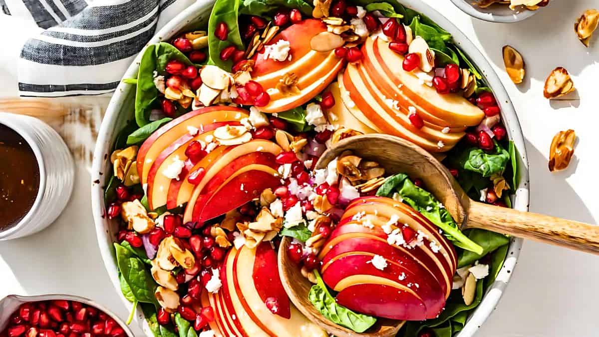 A vibrant salad in a bowl featuring sliced apples, spinach, pomegranate seeds, toasted almonds, and crumbled feta cheese. A wooden spoon rests in the bowl, with a striped towel and small bowls of dressing and toppings nearby.