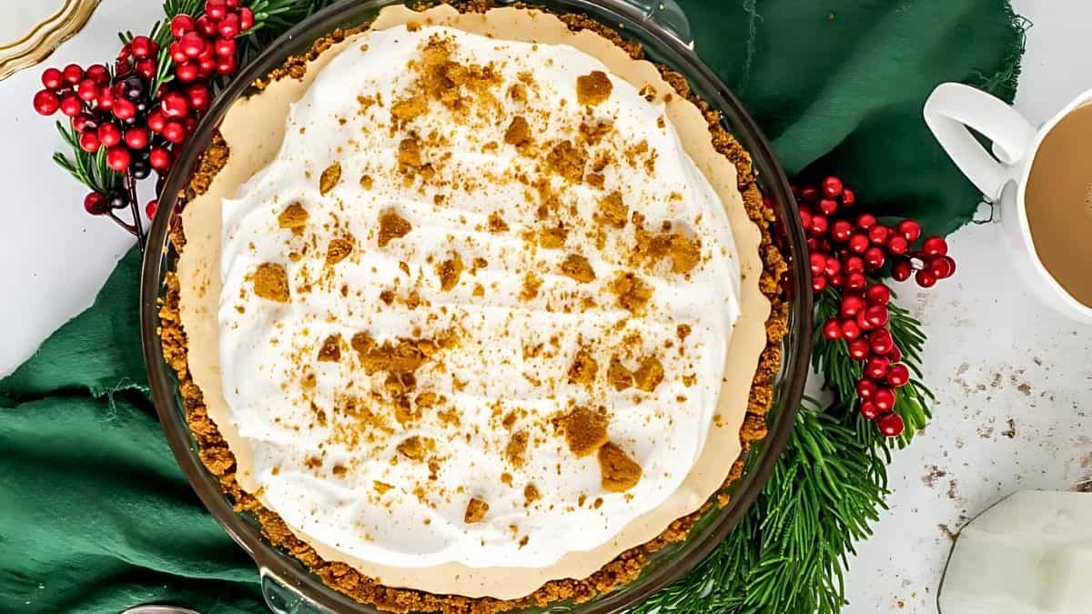 A pie topped with whipped cream and cookie crumbles sits on a green cloth. Red berries and pine branches surround the pie. A cup of coffee is partially visible to the right.