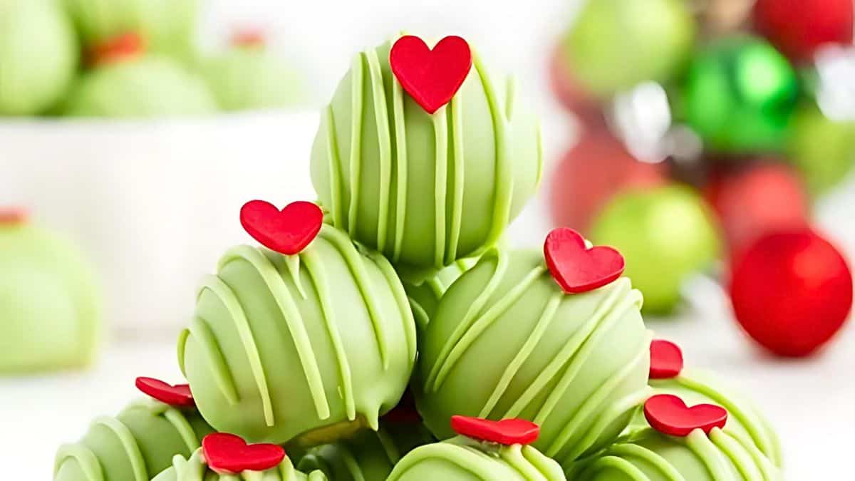 A close-up of green chocolate truffles stacked in a pyramid, drizzled with stripes and topped with small red heart-shaped decorations. Bright, festive ornaments are blurred in the background.