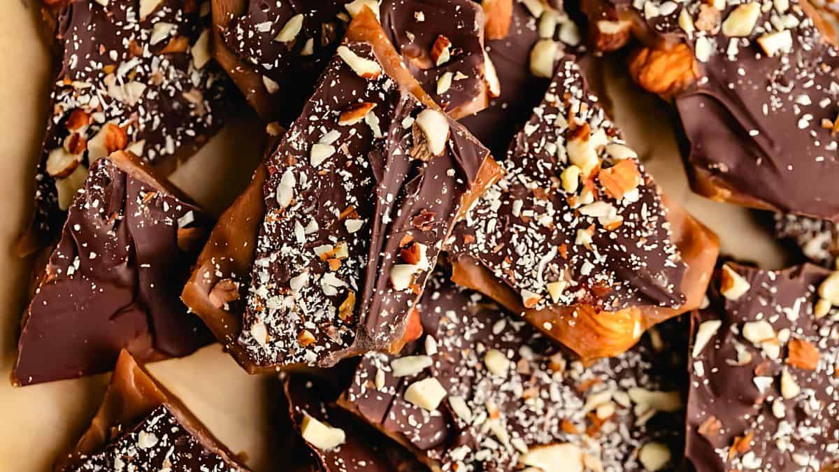 Close-up of homemade toffee pieces covered with a layer of dark chocolate and sprinkled with chopped nuts. The texture of the toffee and glossy chocolate contrasts with the crunchy nut topping.