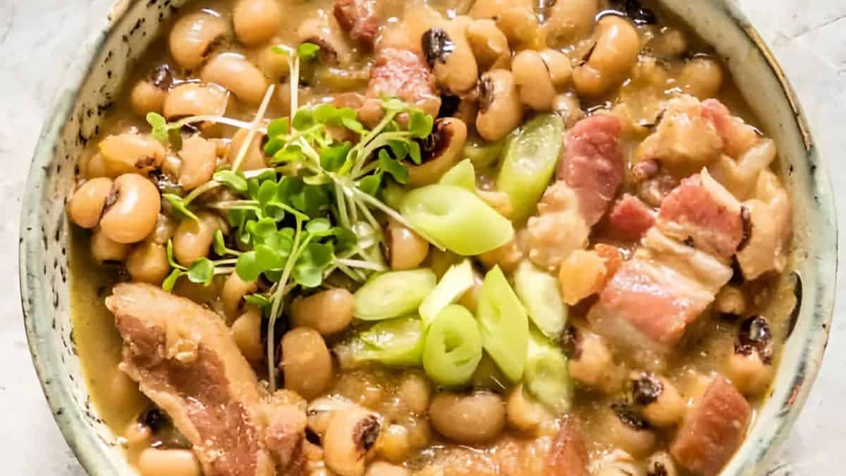 A bowl of black-eyed peas with pieces of bacon, garnished with fresh microgreens and sliced green onions. The creamy beans are in a savory broth, creating a hearty and comforting dish.