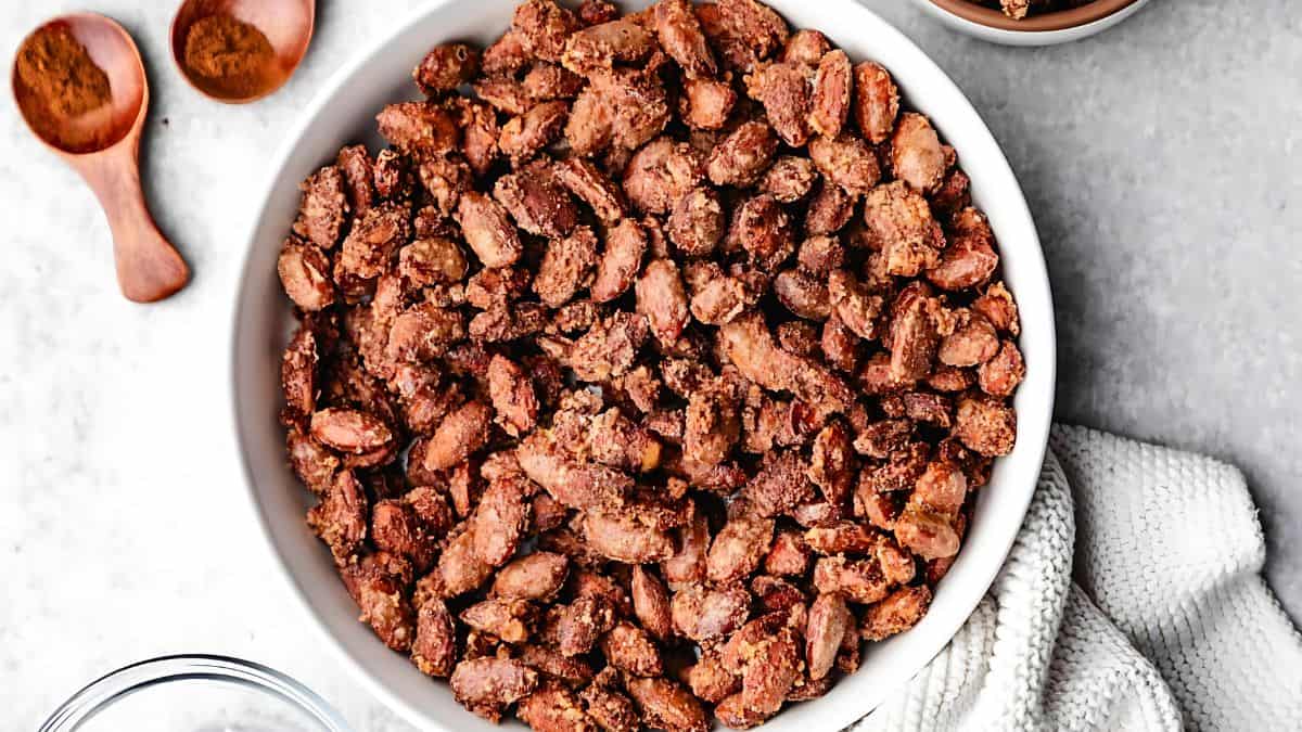A bowl of roasted nuts coated with a spiced seasoning. Two wooden spoons with brown spices are beside it, and a white cloth is partially visible on the side. The surface is a light, speckled gray.