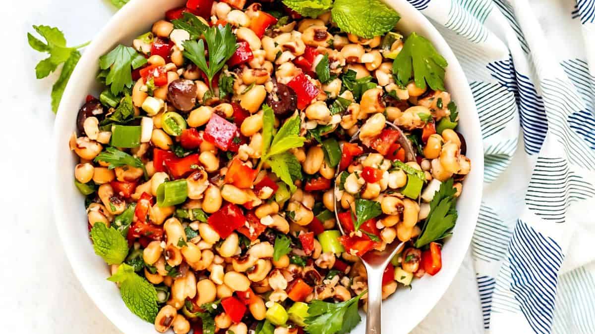 A colorful bean salad in a white bowl, featuring black-eyed peas, chopped red and green bell peppers, sliced green onions, and garnished with fresh parsley and mint leaves. A fork is placed on the side of the bowl.
