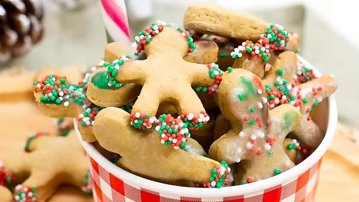 A bowl filled with gingerbread cookies, shaped like people, decorated with red, green, and white sprinkles. The bowl has a red checkered pattern, and a candy cane sticks out from it.
