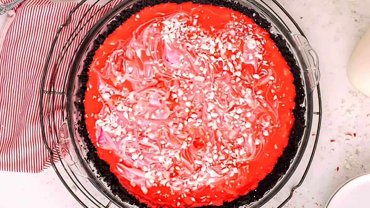 A red and white swirled dessert sits in a chocolate crust, topped with crushed peppermint candy bits. The pie is placed on a cooling rack next to a red-striped towel on a light-colored countertop.
