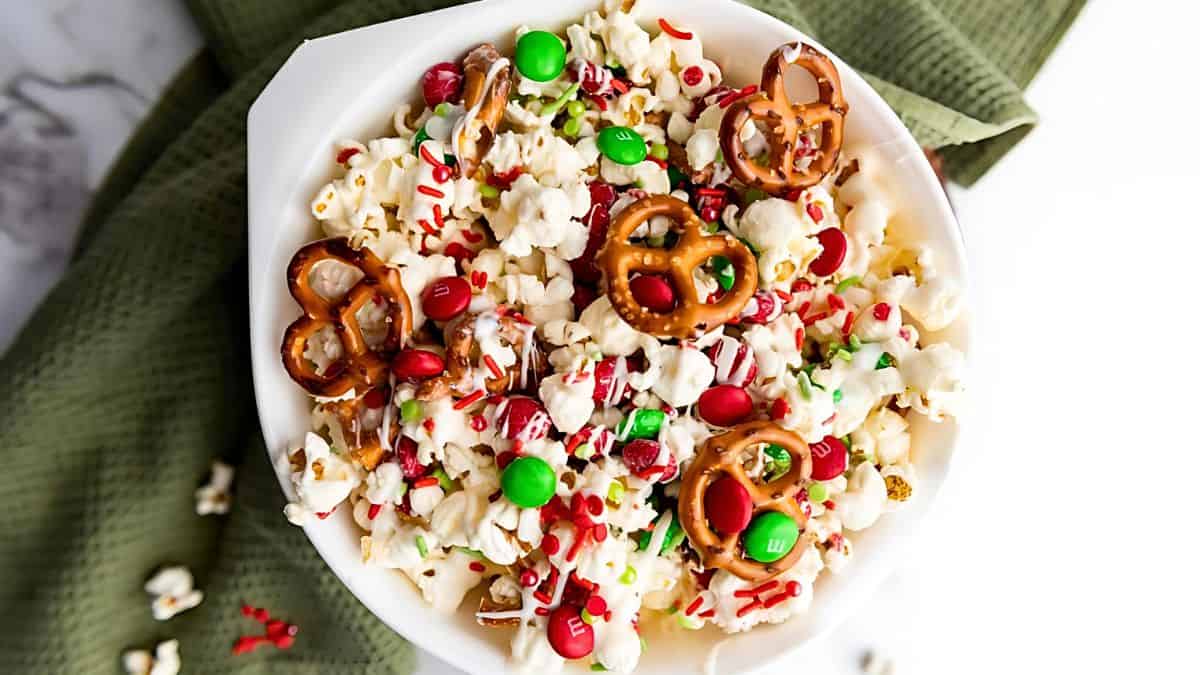 A bowl of festive popcorn topped with red and green candies, pretzel twists, and colorful sprinkles, set on a green textured cloth.