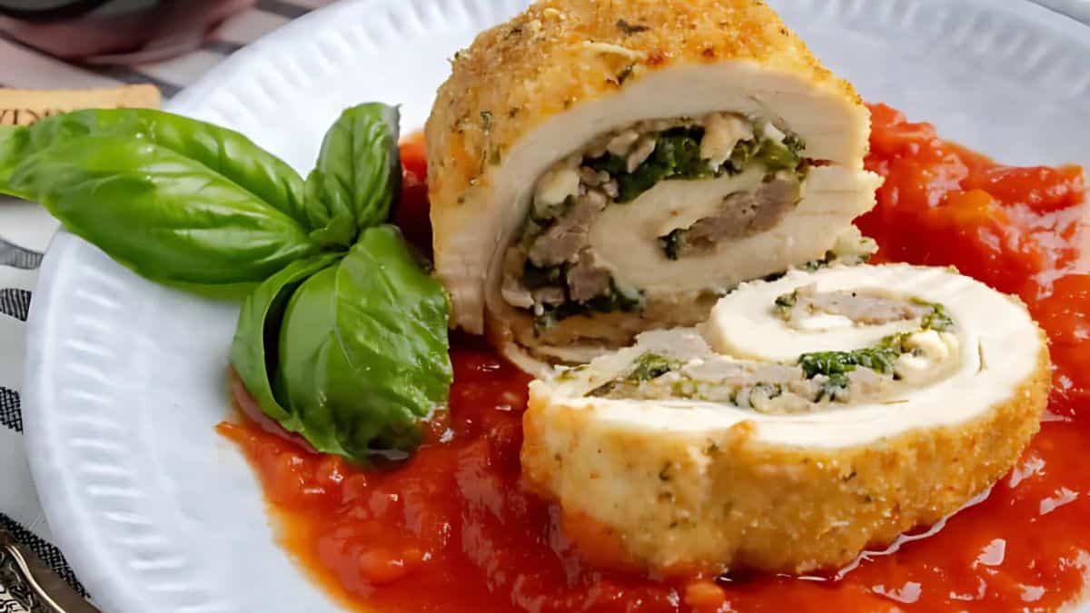 A plated rolled chicken dish stuffed with spinach and cheese, topped with tomato sauce. Fresh basil leaves are garnishing the white plate, adding a touch of green.