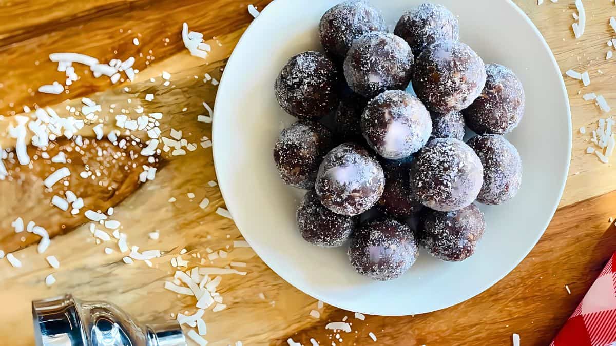 A plate of sugar-coated chocolate balls on a wooden table. Coconut flakes are scattered around, and a silver shaker is nearby, along with a red and white checkered cloth.
