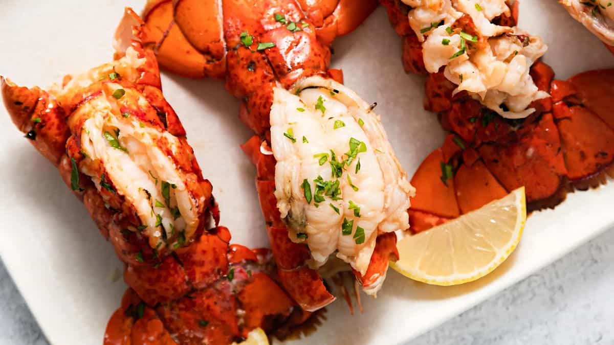 Close-up of cooked lobster tails garnished with parsley, arranged on a white plate with a lemon wedge. The lobster is split to reveal the succulent white meat inside its bright red shell.