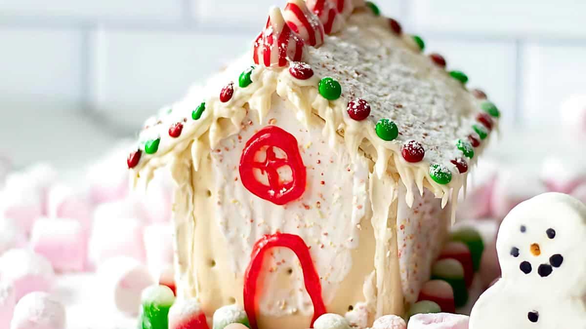 A festive gingerbread house decorated with white icing, red and green candy accents, and a red icing door and window. It is surrounded by pink and white marshmallows, with a small snowman figure on the side.