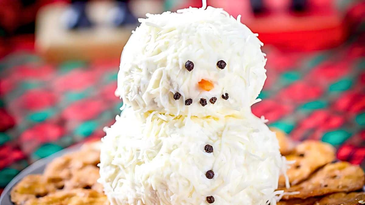 A snowman-shaped cheese ball coated in white cheese shreds, with a carrot nose and small black eyes, sits on a plate surrounded by crackers. The background features vibrant red and green holiday decor.