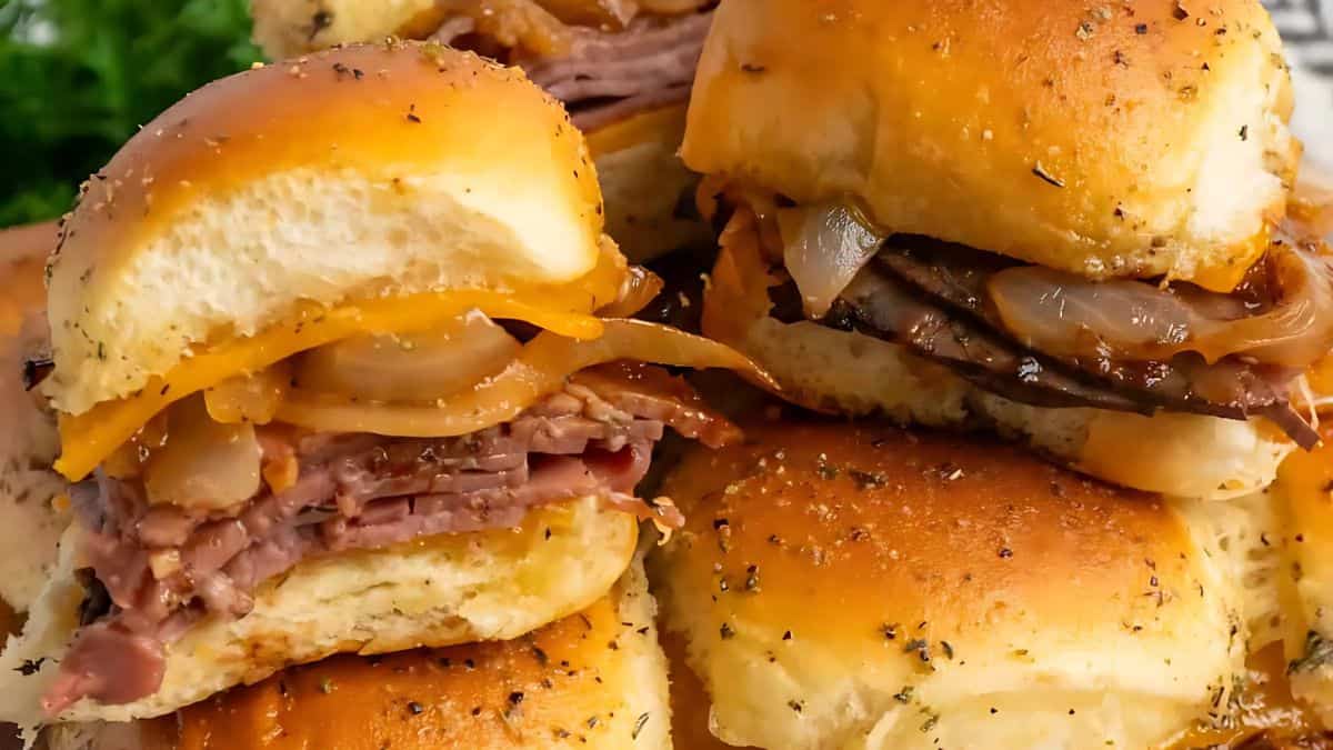 A close-up of a stack of savory sliders filled with sliced roast beef, cheddar cheese, and caramelized onions. The golden-brown buns are sprinkled with herbs. The background is slightly blurred with hints of green leaves.