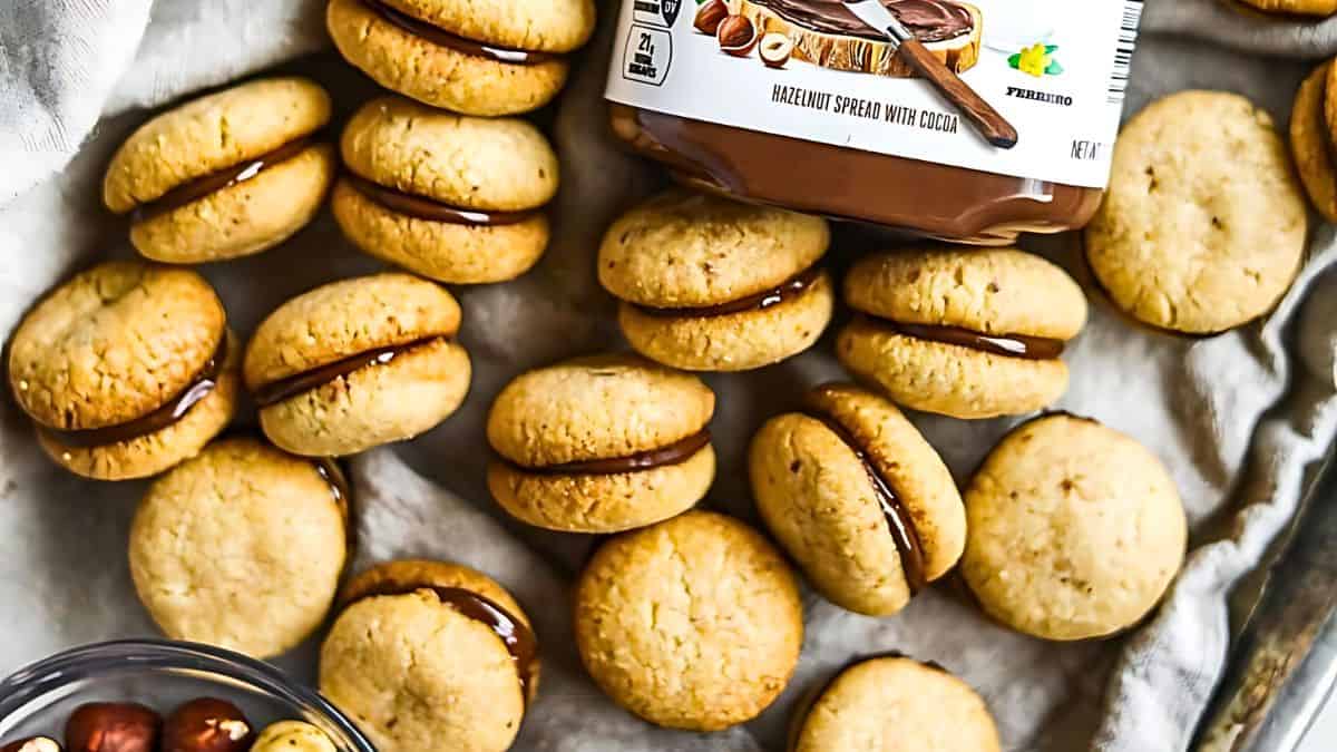 A collection of sandwich cookies filled with chocolate hazelnut spread is scattered on a cloth. A jar of hazelnut spread is placed on top of some cookies, and a small bowl of hazelnuts is visible in the corner.