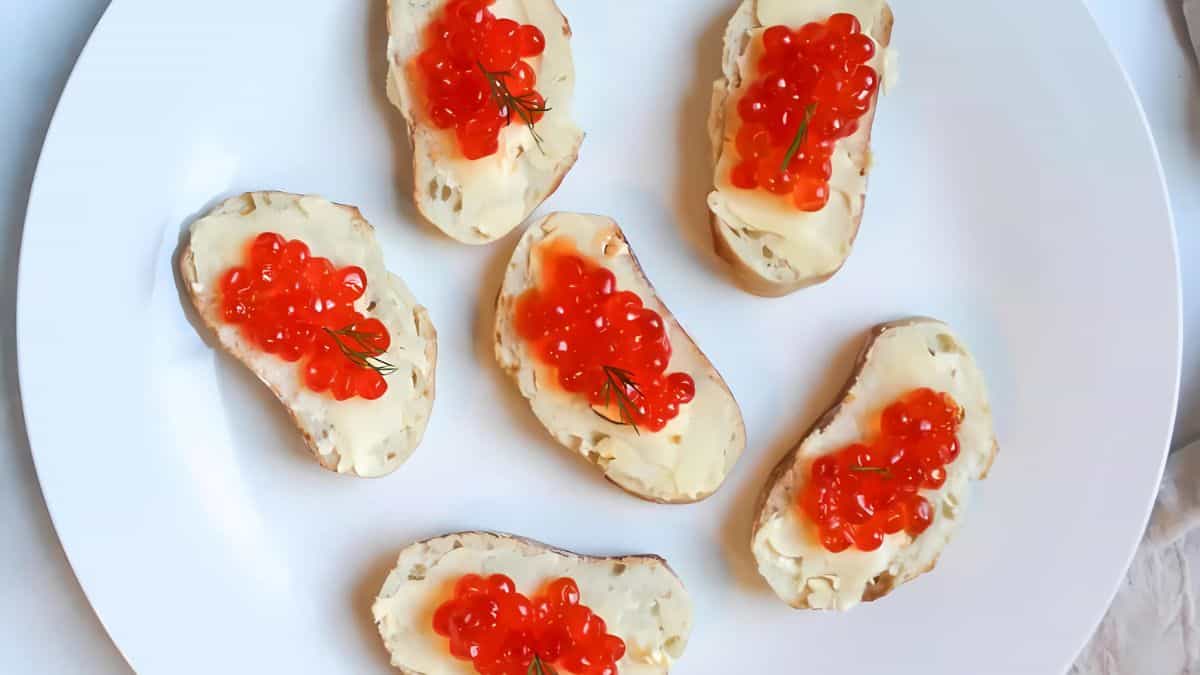 A white plate with six slices of bread topped with butter and red caviar, garnished with small sprigs of dill.