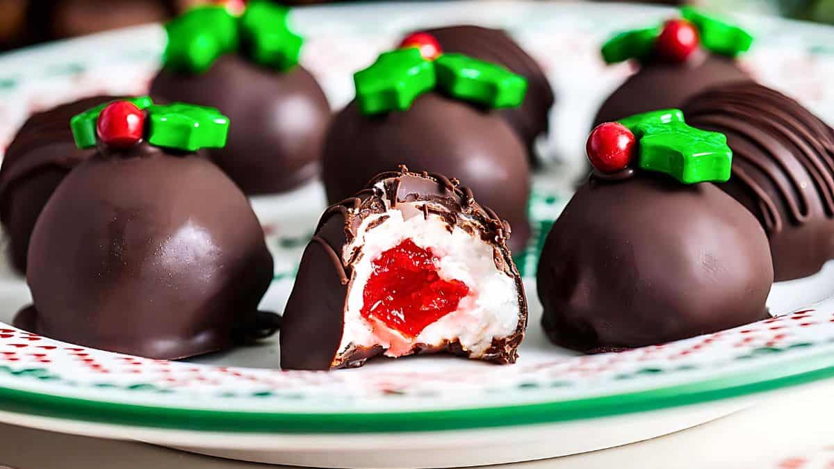 A plate of chocolate truffles, each topped with a decorative green icing shaped like holly leaves and a red candy berry. One truffle is cut open, revealing a creamy white filling and a vibrant red center.