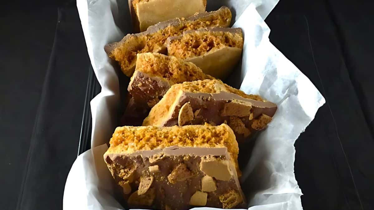 Slices of honeycomb chocolate bars arranged in a paper-lined basket on a dark surface. The bars feature a layer of golden honeycomb topped with chocolate, creating a crunchy and sweet treat.