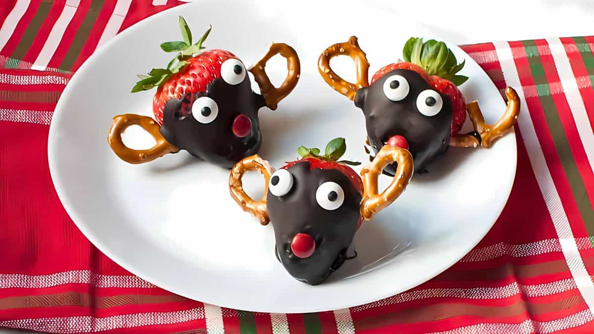 Three chocolate-covered strawberries decorated as reindeer with pretzel antlers, candy eyes, and red noses are arranged on a white plate. The plate is on a red and green plaid tablecloth.
