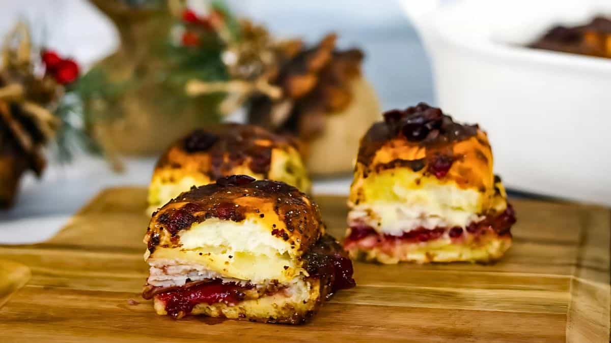 Three turkey slider sandwiches with cranberry sauce on a wooden board, garnished with seeds. A blurred festive background with pinecones and holiday decor is visible.