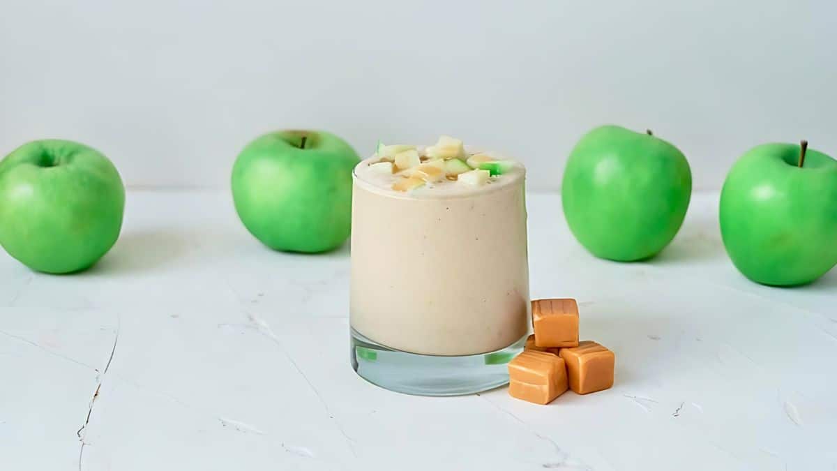 A creamy caramel apple smoothie in a glass topped with chopped apple pieces. Four green apples and four caramel candies are arranged beside it on a white surface.