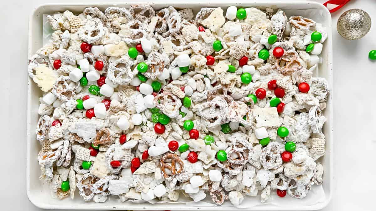 A tray filled with a festive snack mix, including pretzels, cereal pieces, mini marshmallows, red and green candies, and white chocolate-coated treats. The mix is dusted with powdered sugar, and a silver ornament is visible beside the tray.