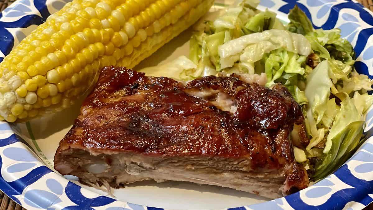 A plate with a serving of glazed barbecue ribs, a cob of corn, and a side of cabbage salad, all on a decorative blue and white paper plate.