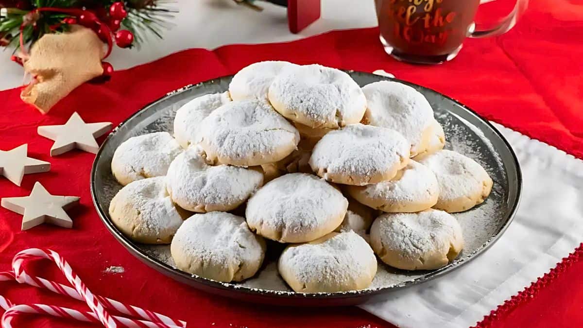 A plate of powdered sugar-covered cookies is arranged in a pyramid shape on a table. Nearby, there are red and white decorations, including candy canes, white star ornaments, and a festive mug. The setting conveys a holiday theme.