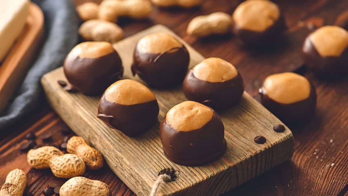 A wooden board with several chocolate-covered peanut butter balls, resembling buckeye candies. Whole peanuts are scattered on the wooden surface around the board. The background features a mix of brown and warm tones.