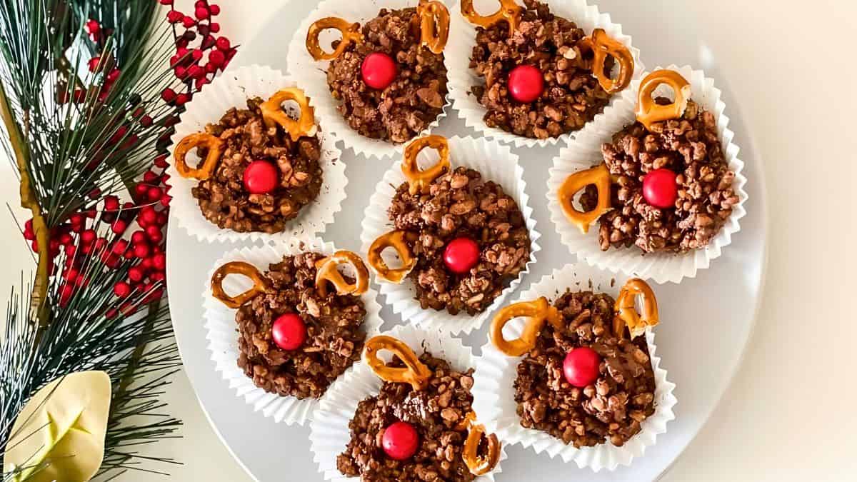 A plate of eight chocolate treats decorated with pretzel antlers and red candy noses, arranged to resemble reindeer. They're placed on white paper liners. Pine branches and red berries are on the left side of the image.