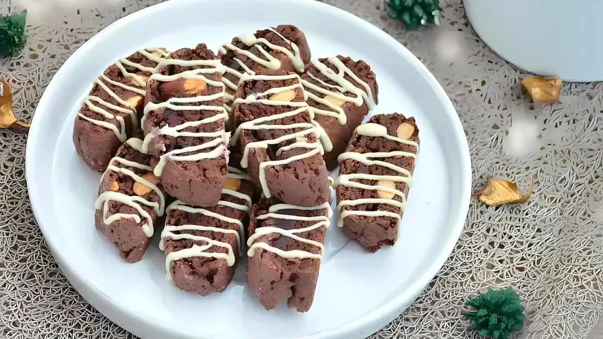 A white plate holds a batch of chocolate biscotti drizzled with white icing. The biscotti are arranged neatly, showcasing their almond pieces. The background features a festive setting with small decorative elements.