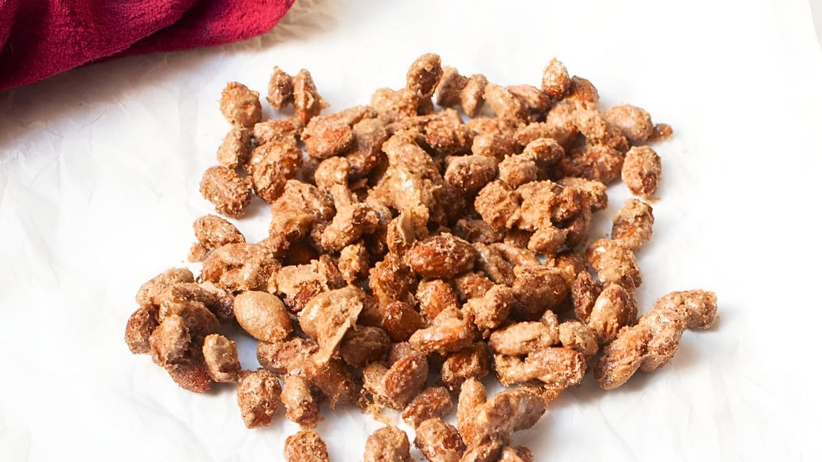 A pile of candied almonds is spread out on a white surface. The almonds are coated with a crystallized sugary layer, giving them a shiny, textured appearance. A red cloth is partially visible in the corner.