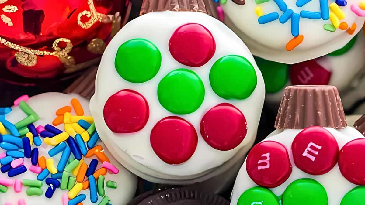 A close-up of decorated holiday cookies. One cookie has red and green candies arranged in a circle on white icing. Another cookie is topped with colorful sprinkles. A chocolate candy decorates a cookie in the background.