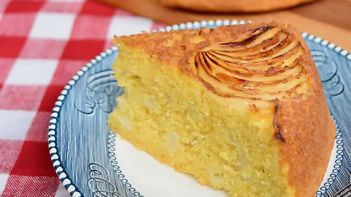 A slice of cornbread cake on a blue and white plate, set on a red and white checkered tablecloth. The cake displays a decorative top and a moist, dense texture.