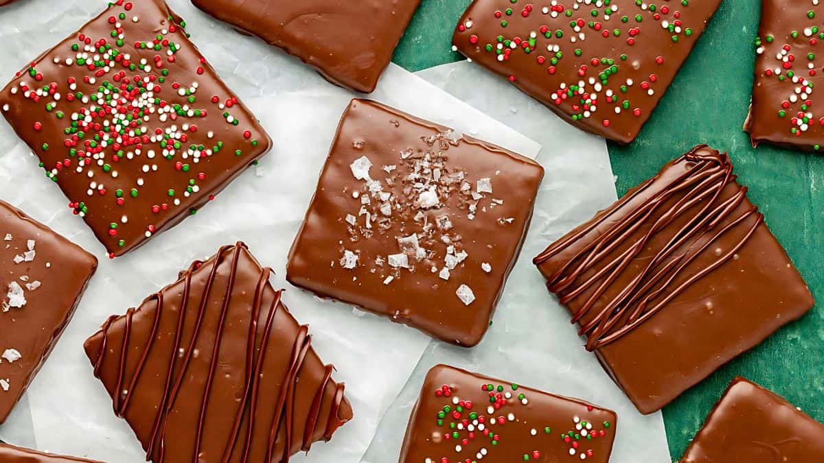 An assortment of square chocolate-covered cookies on parchment paper and a green background. Some are decorated with colorful sprinkles, some with striped drizzle, and one with salt flakes.
