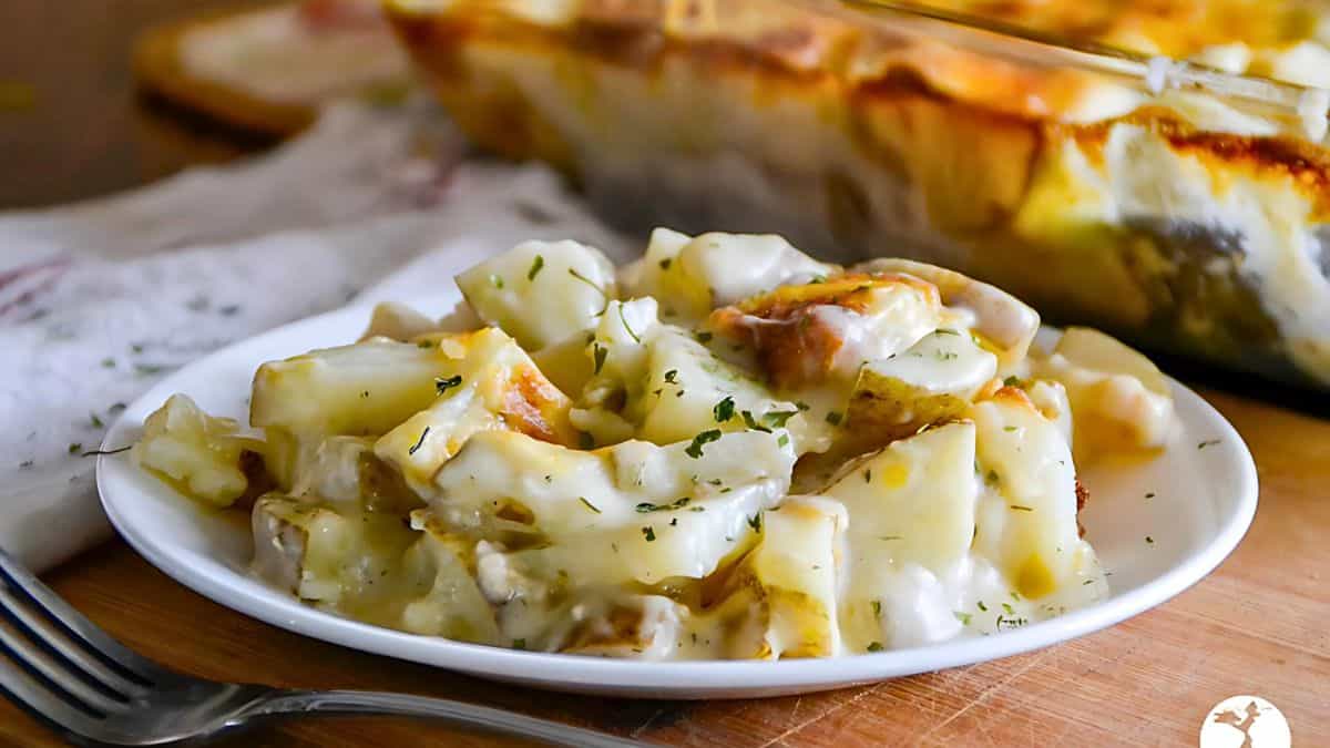 A plate of creamy scalloped potatoes topped with herbs is placed on a wooden surface. In the background, a casserole dish filled with more of the same dish is partially visible, next to a folded napkin and a fork.