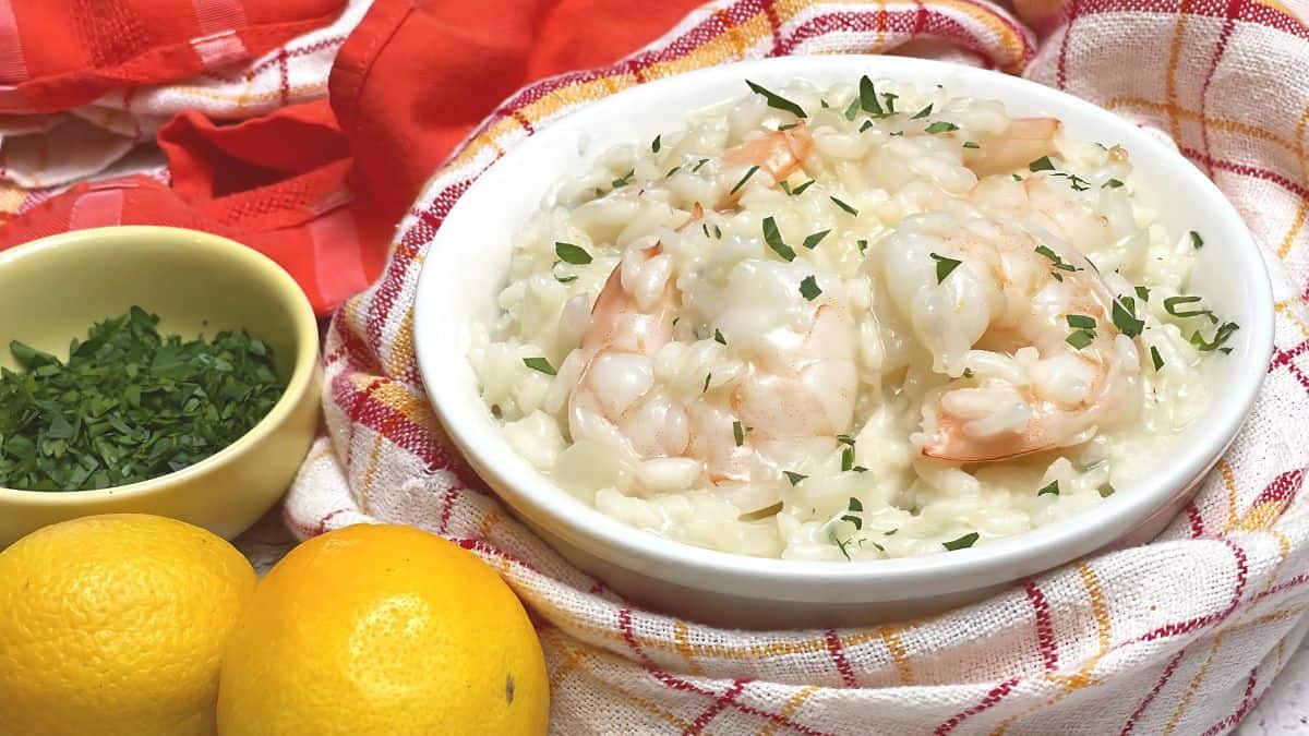 A bowl of creamy shrimp risotto garnished with chopped parsley sits on a red and white checked cloth. Beside it are two lemons and a small bowl of additional parsley.