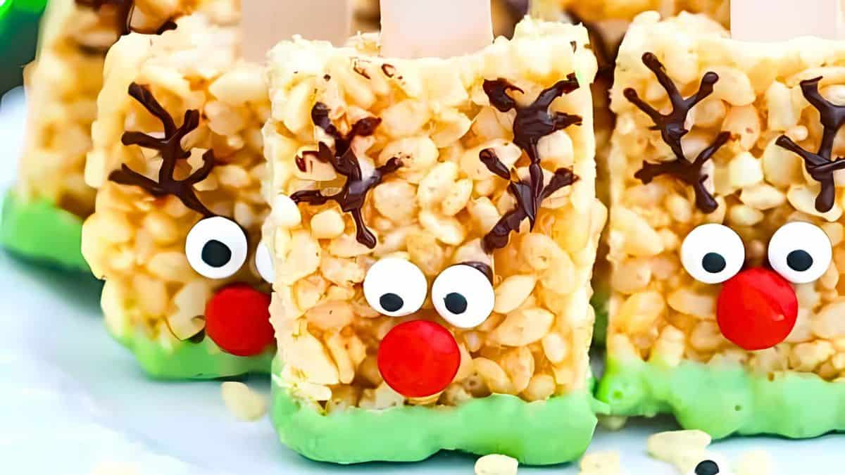 Three reindeer-themed Rice Krispies treats with candy eyes, red candy noses, and chocolate antlers. The bottoms are dipped in green icing, creating a festive appearance.
