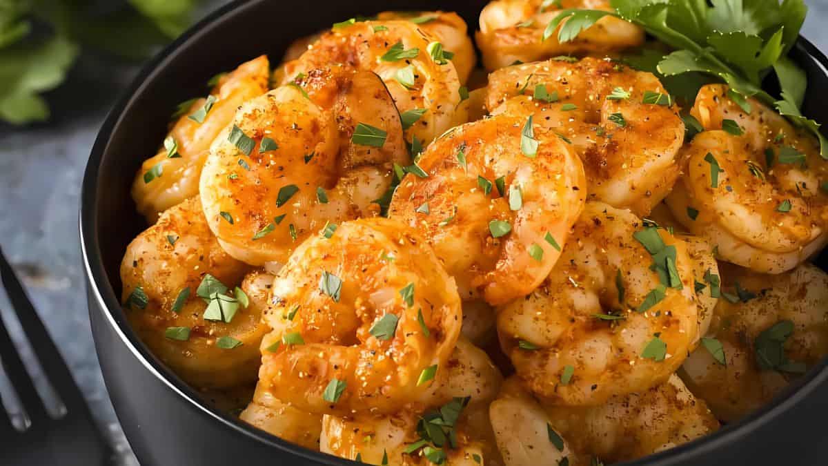 A bowl filled with seasoned shrimp garnished with chopped herbs. The shrimp are golden and arranged neatly, with a sprig of parsley on the side. The background features blurred green leaves and a fork.