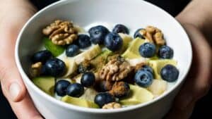 A close-up of a bowl filled with sliced bananas, blueberries, walnuts, and chunks of green fruit, possibly avocado, sprinkled with a brown spice, likely cinnamon. The bowl is being held in two hands against a dark background.
