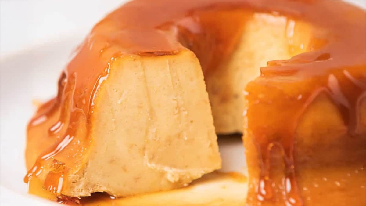 A close-up of creamy cheese flan with a slice cut out, showing its smooth texture and glossy caramel sauce dripping down the sides.